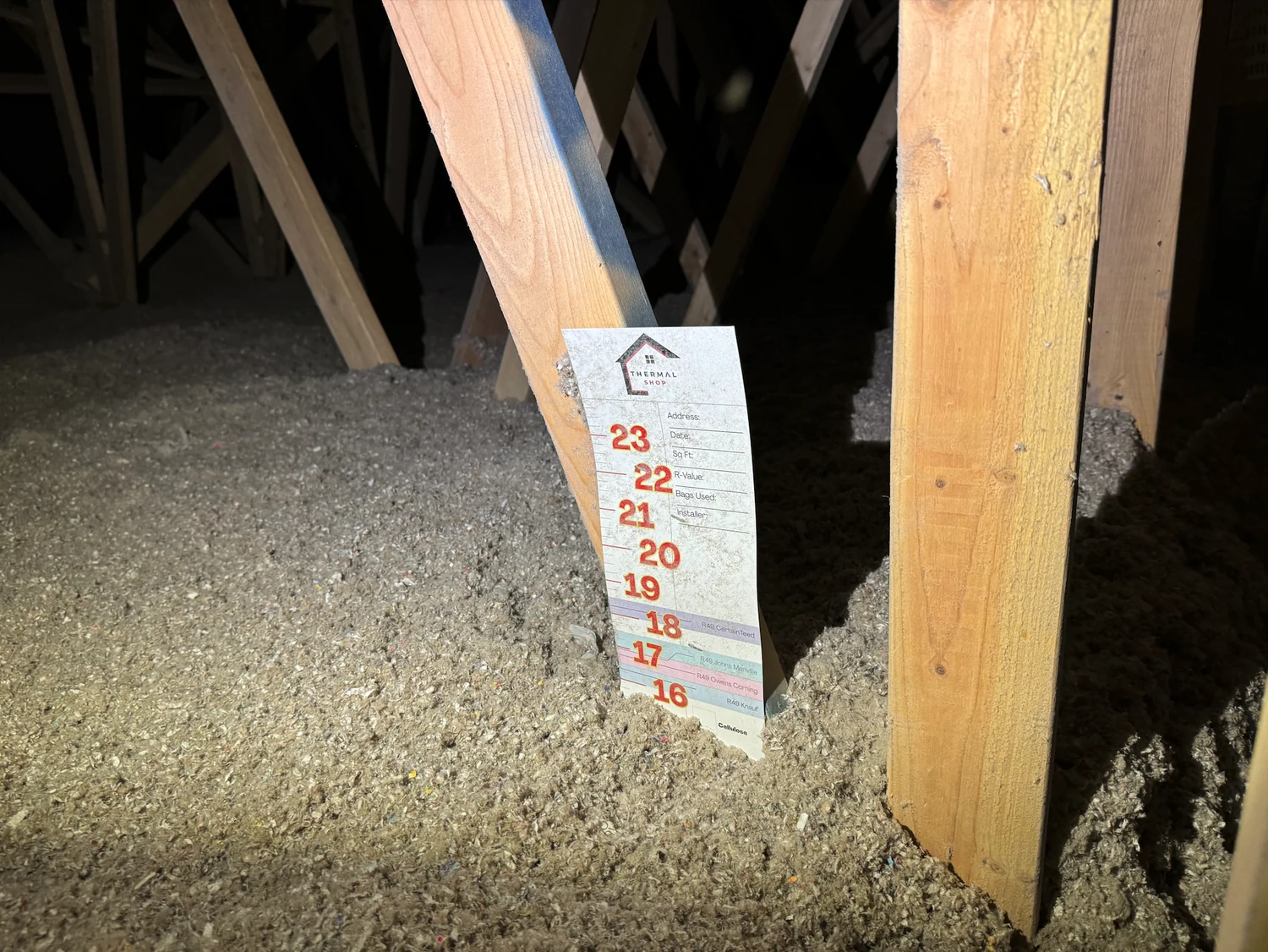 Finished attic floor of grey loose fill with a depth marker card beside a truss