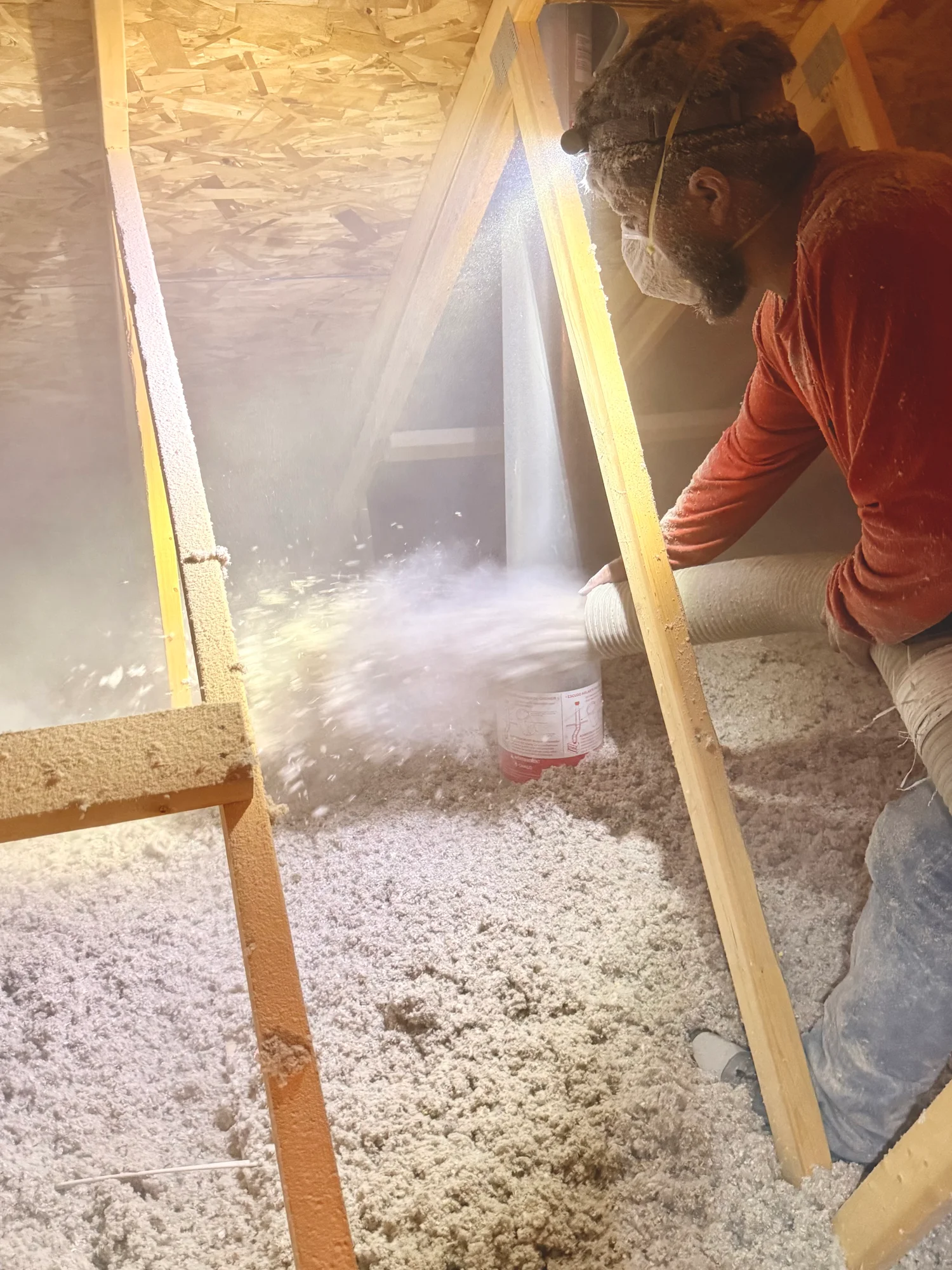 Installer blowing loose-fill insulation across an attic floor, lit by a work light between the trusses
