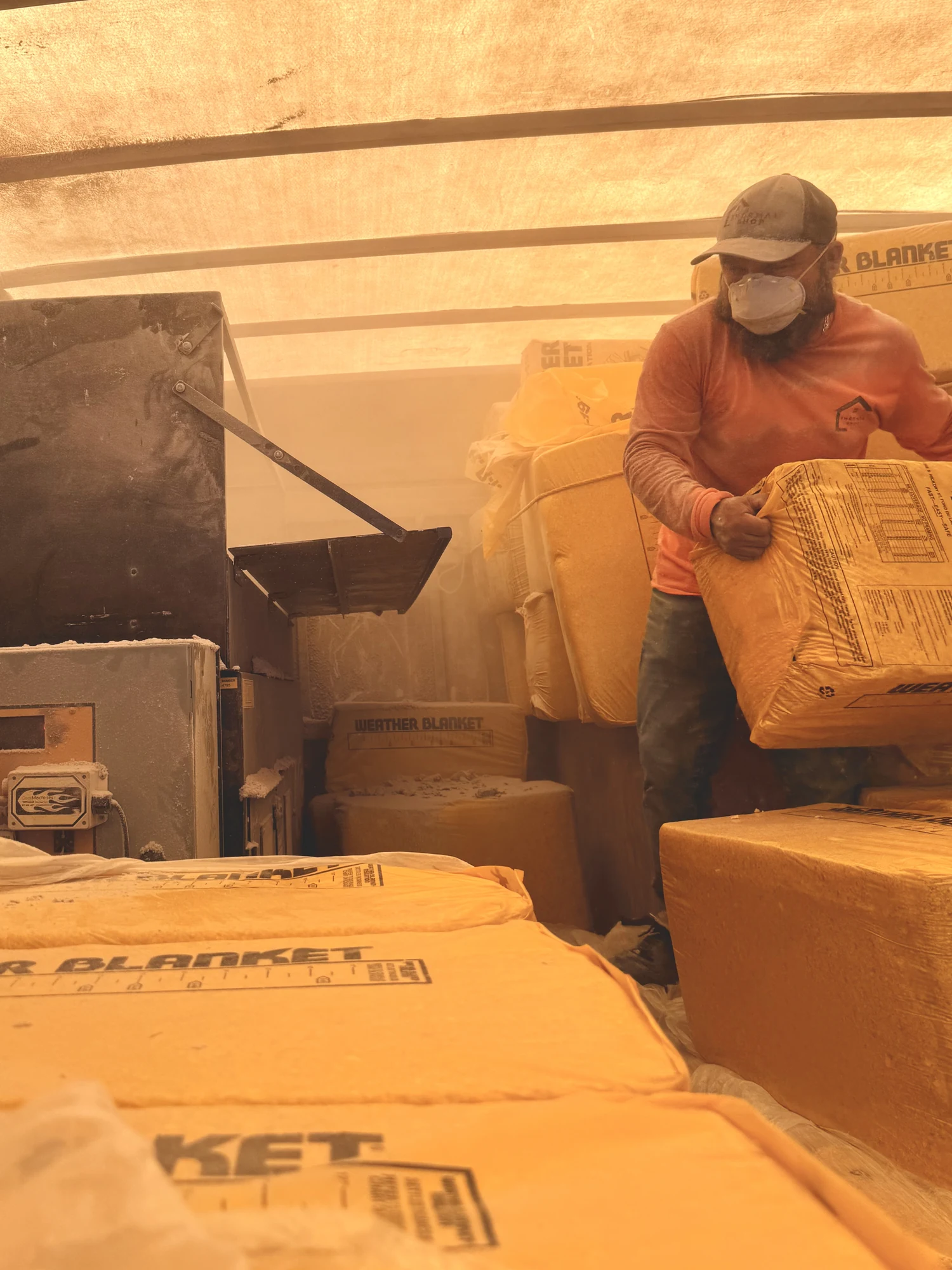 Installer in a respirator and cap lifting a bale of insulation into the blowing machine in the back of a dust-filled box truck