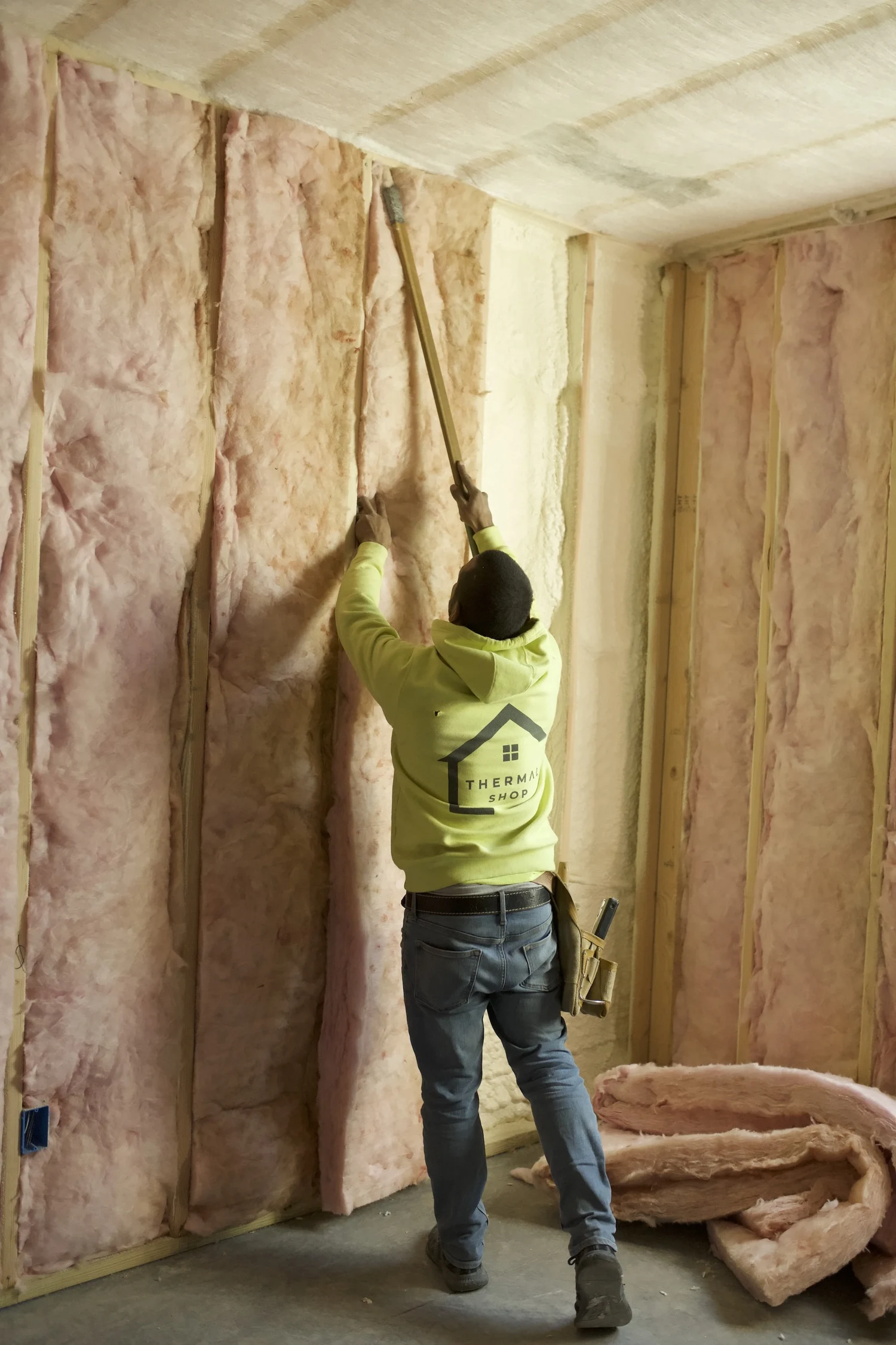 Installer in a high-visibility hoodie pressing a fiberglass batt into a stud cavity
