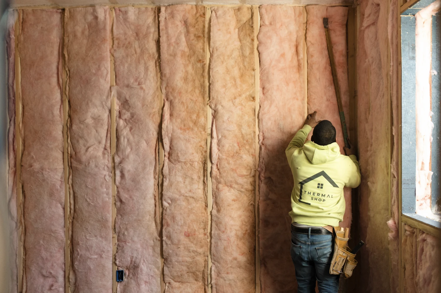 Thermal Shop installer in a company hoodie pressing fiberglass batts into a stud wall