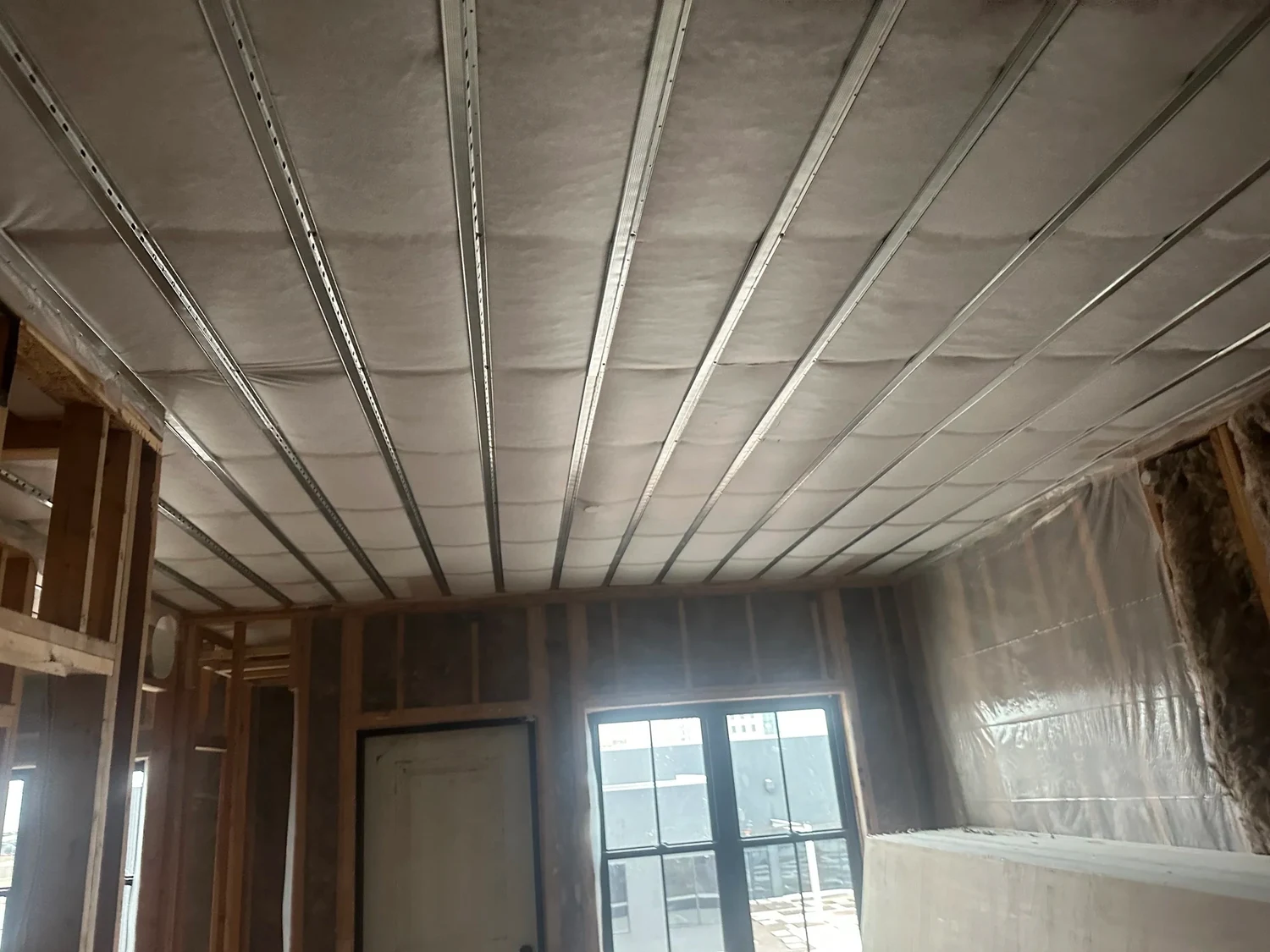 Netted, blown ceiling crossed by metal furring channels above a framed room with a window