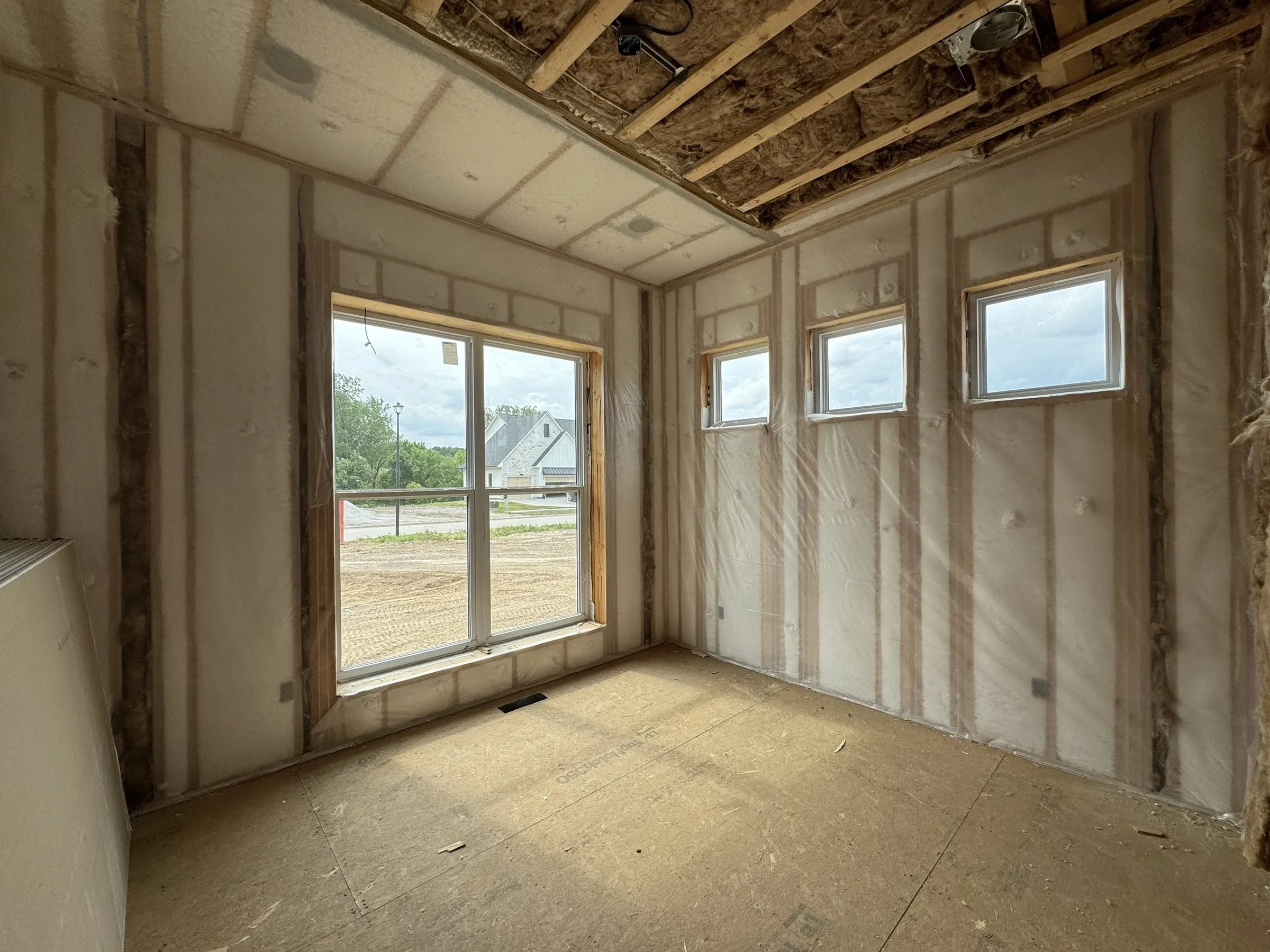 A new-home room with walls and ceiling insulated behind netting, swept and ready for drywall, with the subdivision outside the window