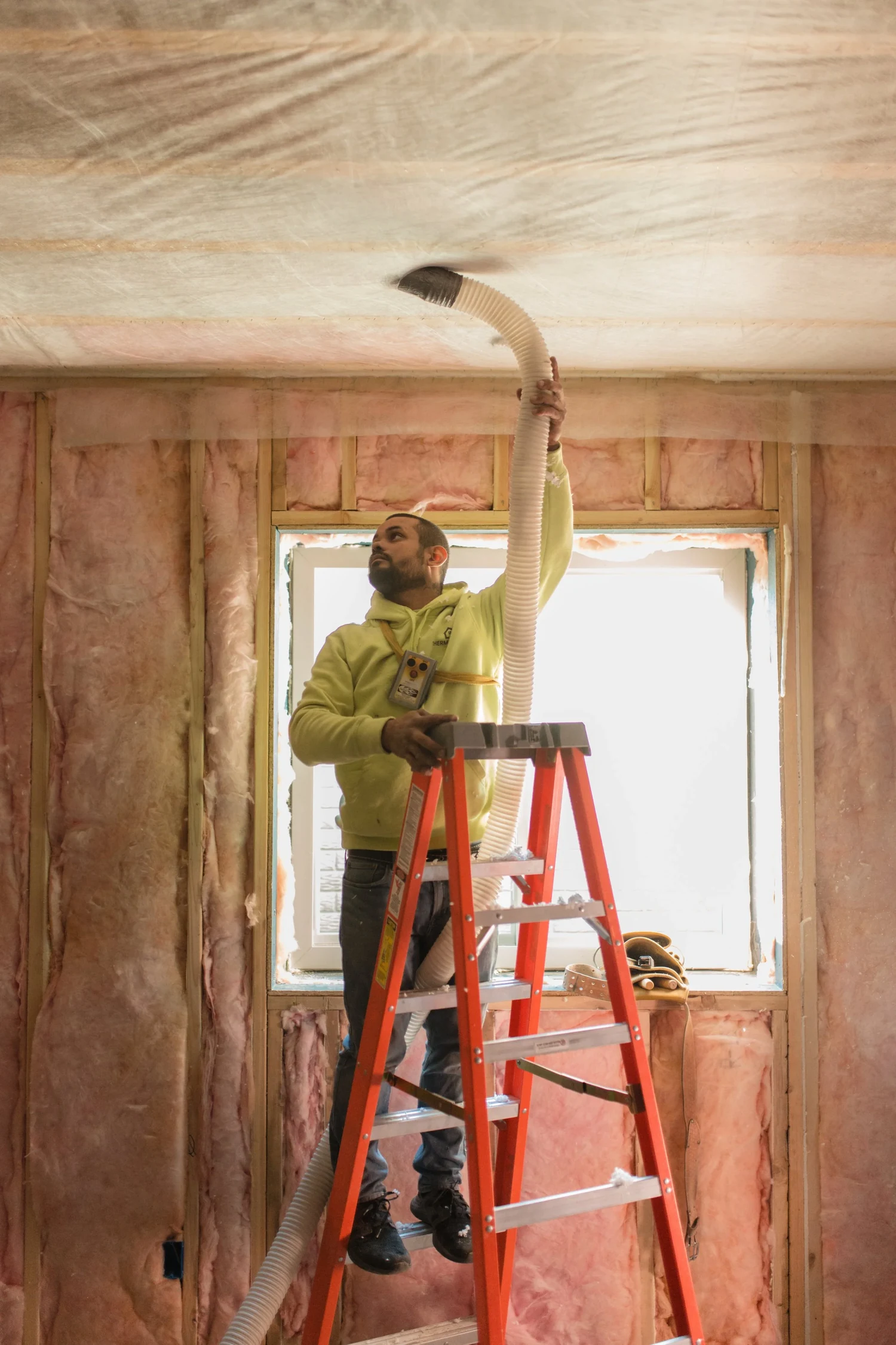 Installer on a red stepladder blowing fiberglass behind the netting on a ceiling, with pink batts in the walls behind him