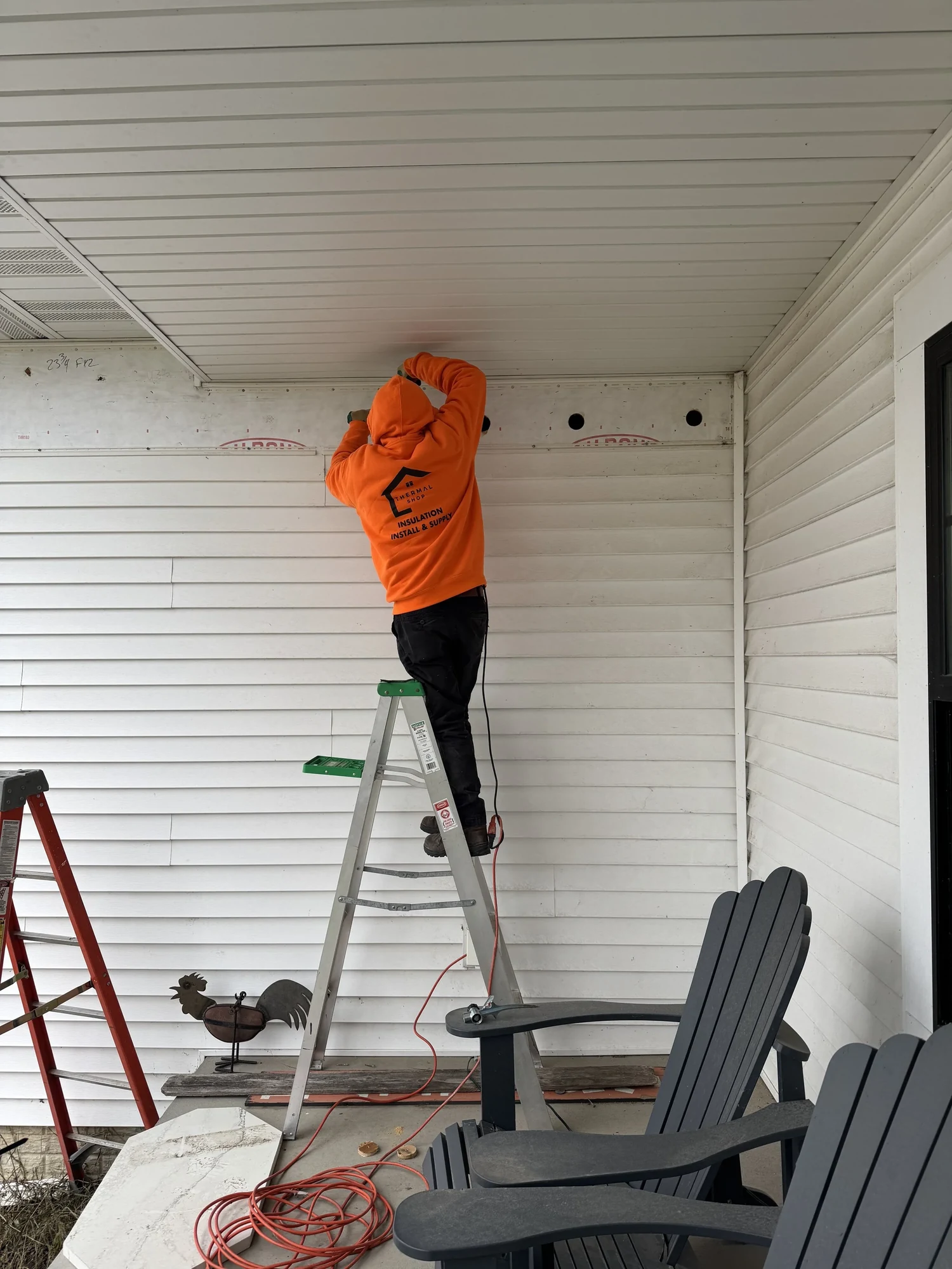 Installer on a stepladder working along a row of openings at the top of a sided wall under the porch