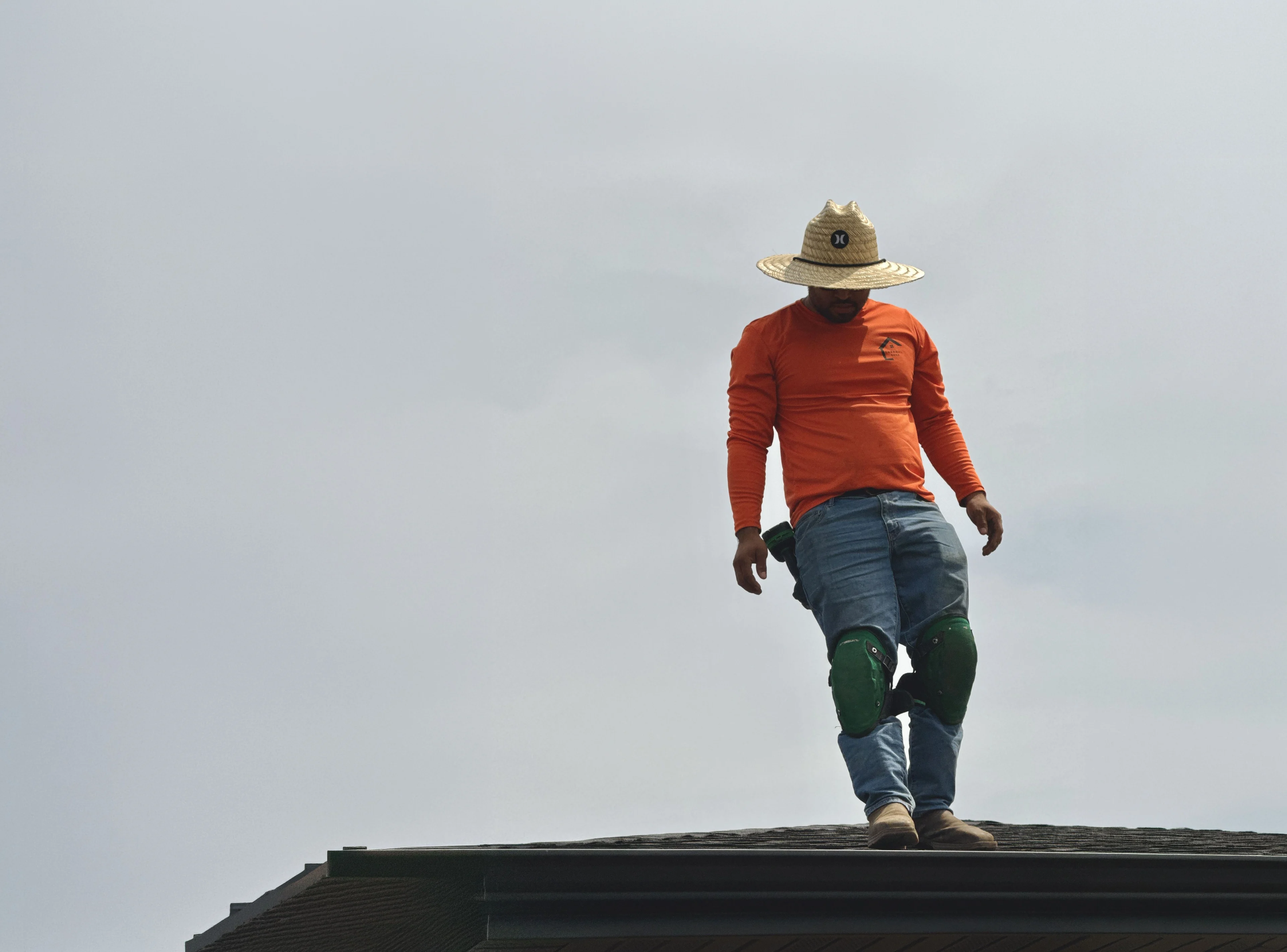Thermal Shop gutter installer in a straw hat, orange shirt, and green knee pads walking along the roof edge above a new gutter