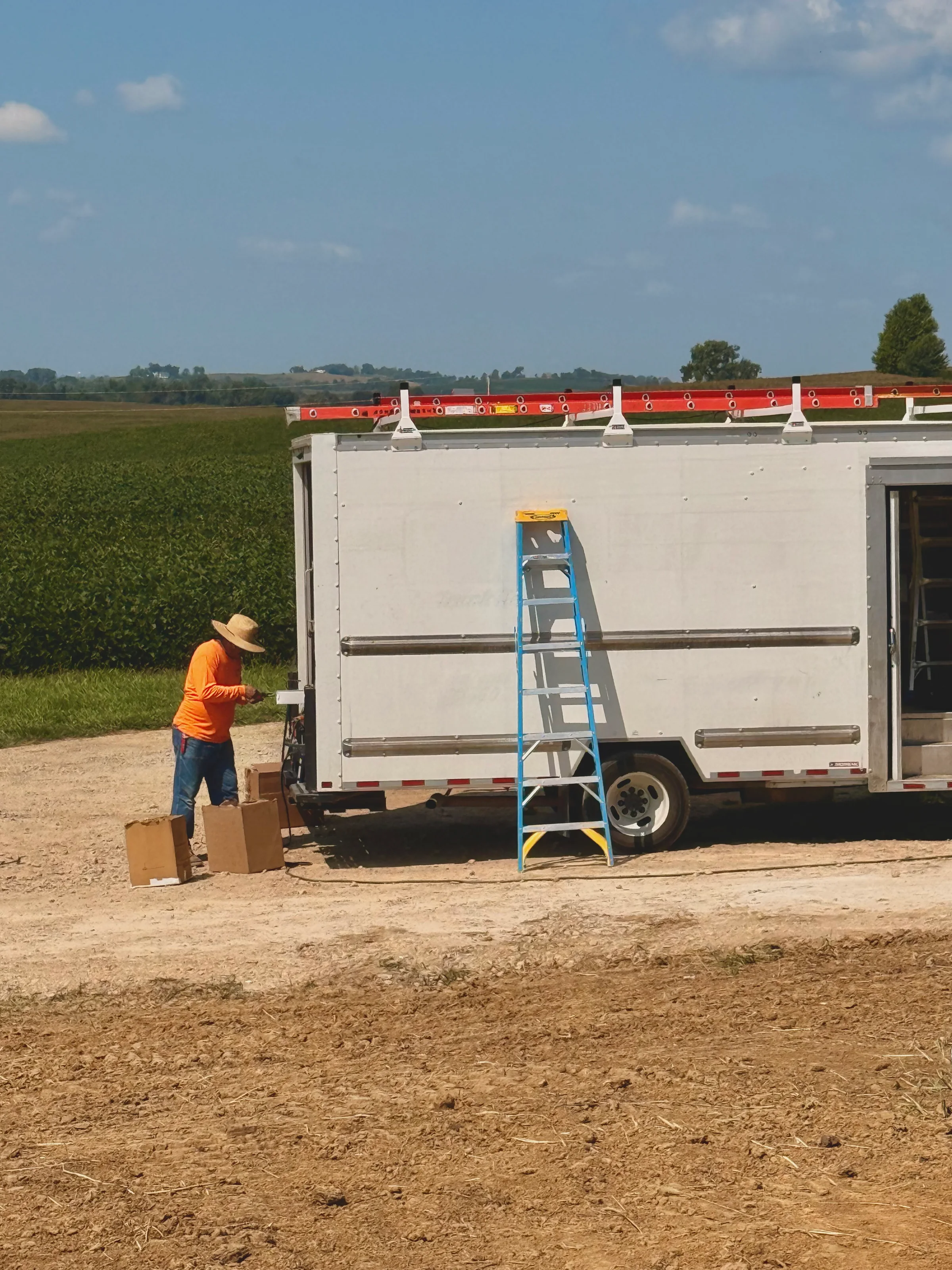 Thermal Shop installer in a hi-vis orange shirt and straw hat working the gutter machine at the back of the white box truck, flat coil cartons on the gravel beside him