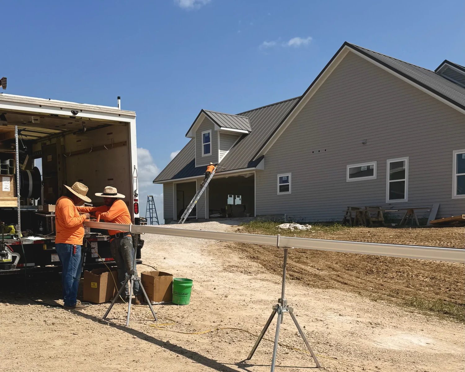 Two Thermal Shop installers at the open back of a box truck, a formed gutter on stands in the yard, and a third on a ladder at a new gray house