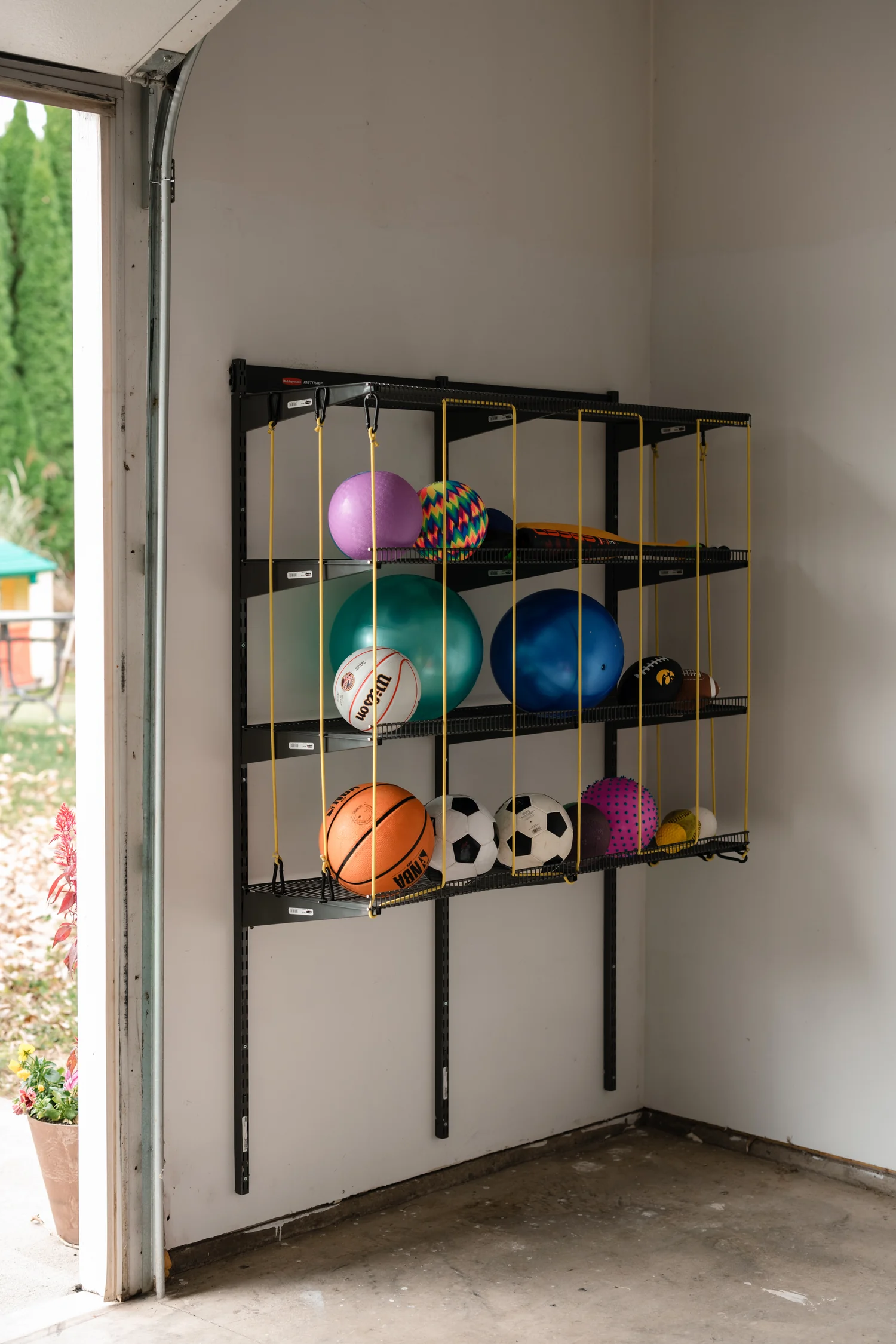Sports balls held behind yellow bungee cords on a gunmetal garage ball rack beside an open door