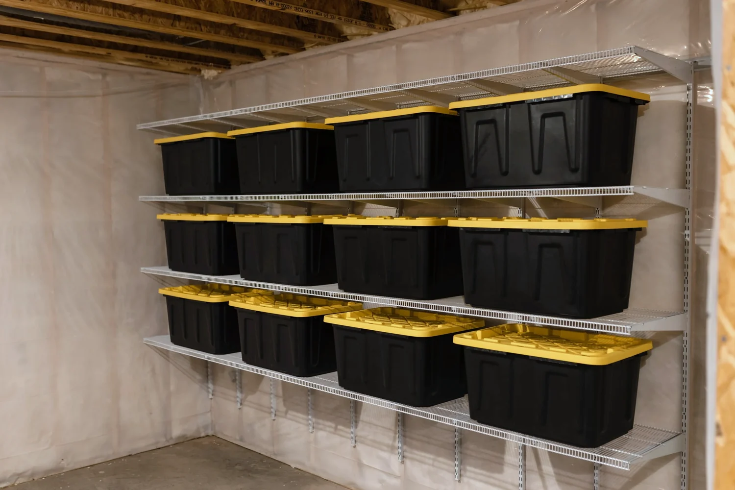 Black storage bins with yellow lids on white wire shelves in an unfinished basement