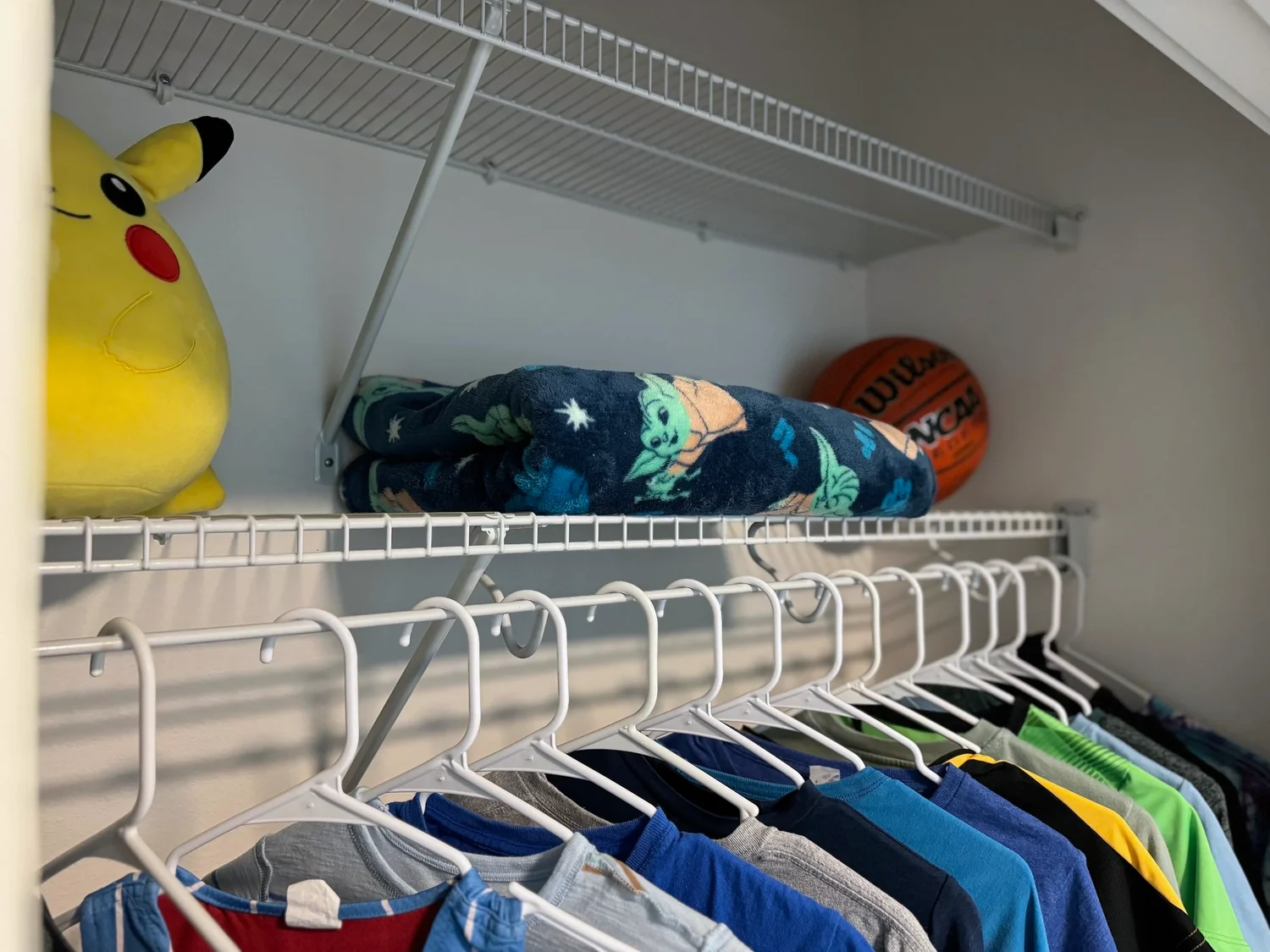Close view of a wire closet shelf holding a stuffed toy and a basketball above hanging shirts