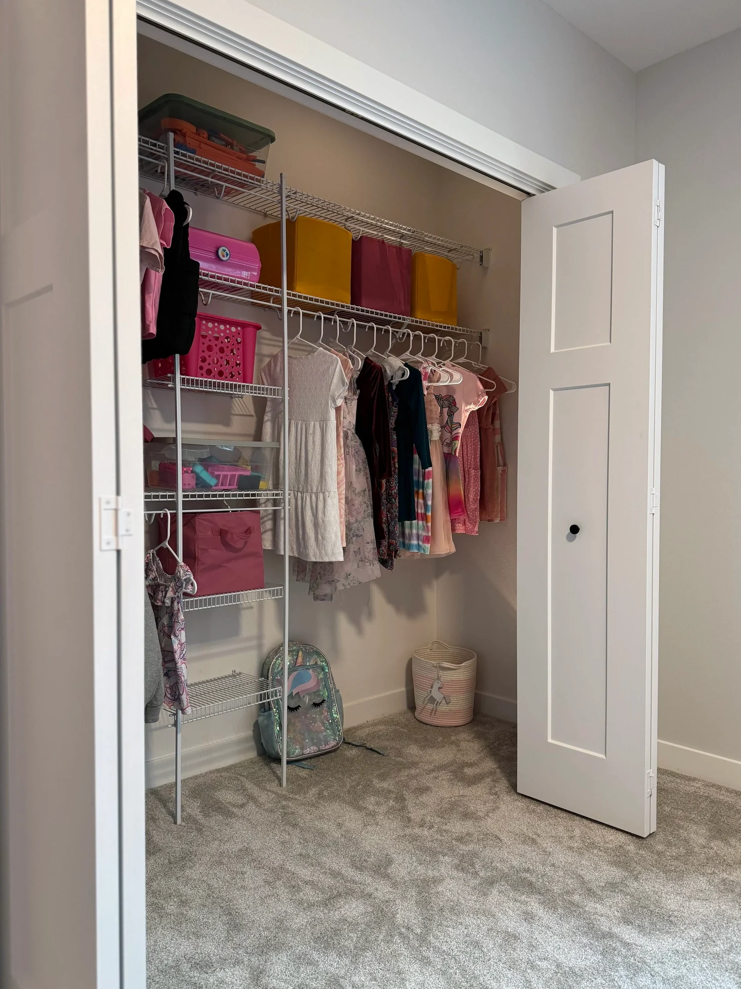 A child's closet with wire shelving, colored bins, and hanging clothes, seen through open doors