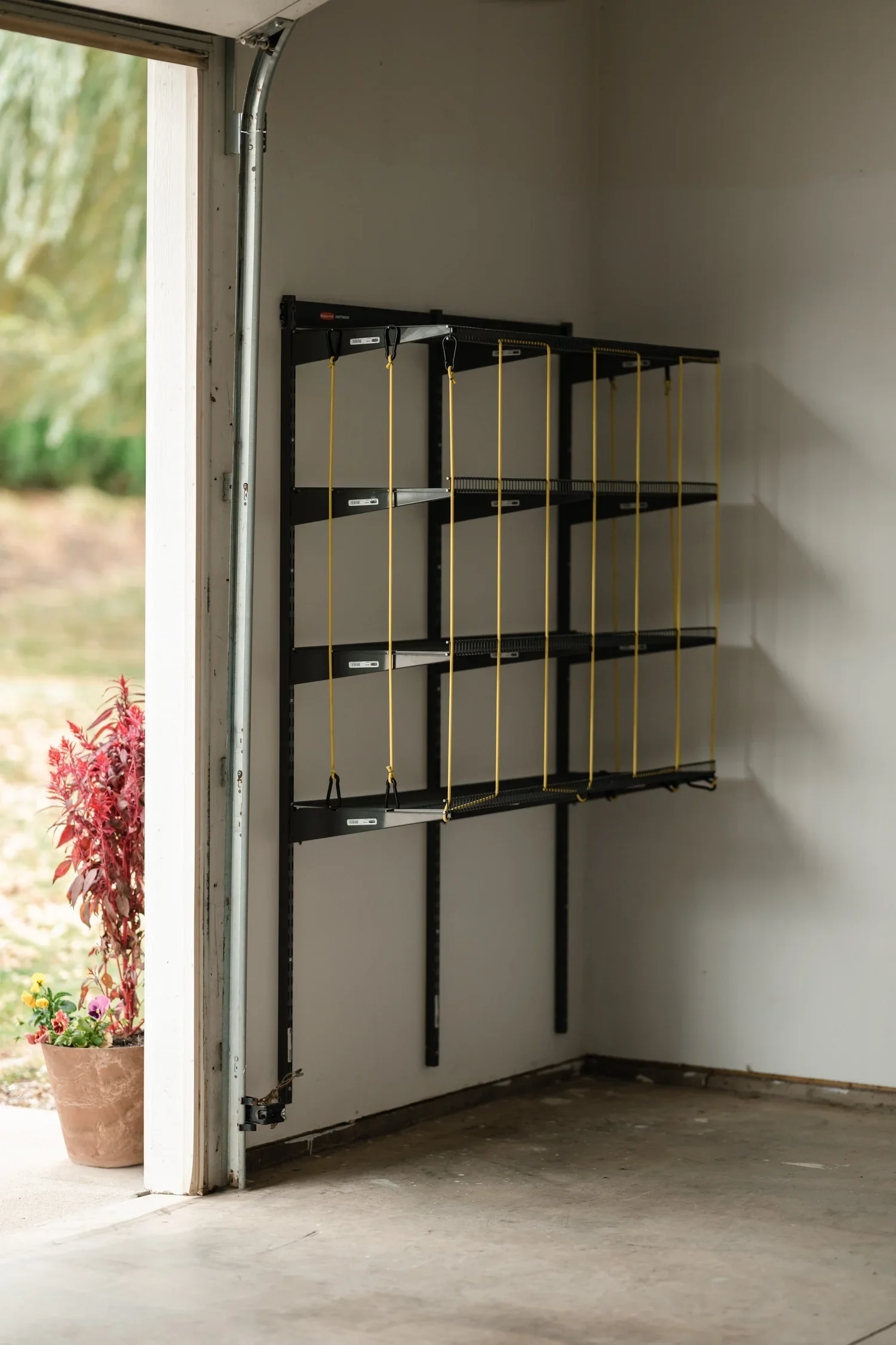 An empty ball rack with yellow bungee cords on gunmetal wall rails, beside an open garage door