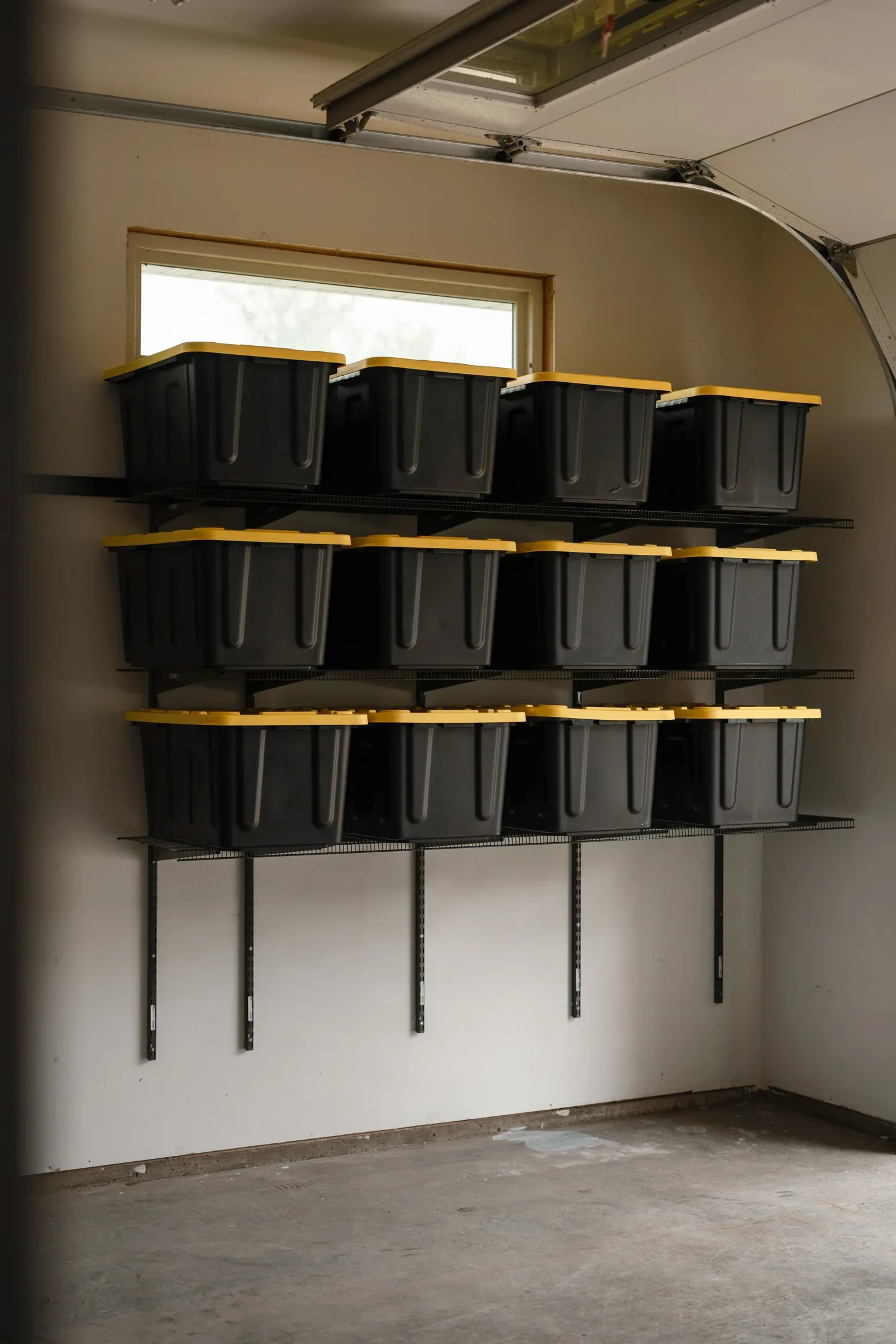 Rows of black storage bins with yellow lids on gunmetal shelves beneath a garage window