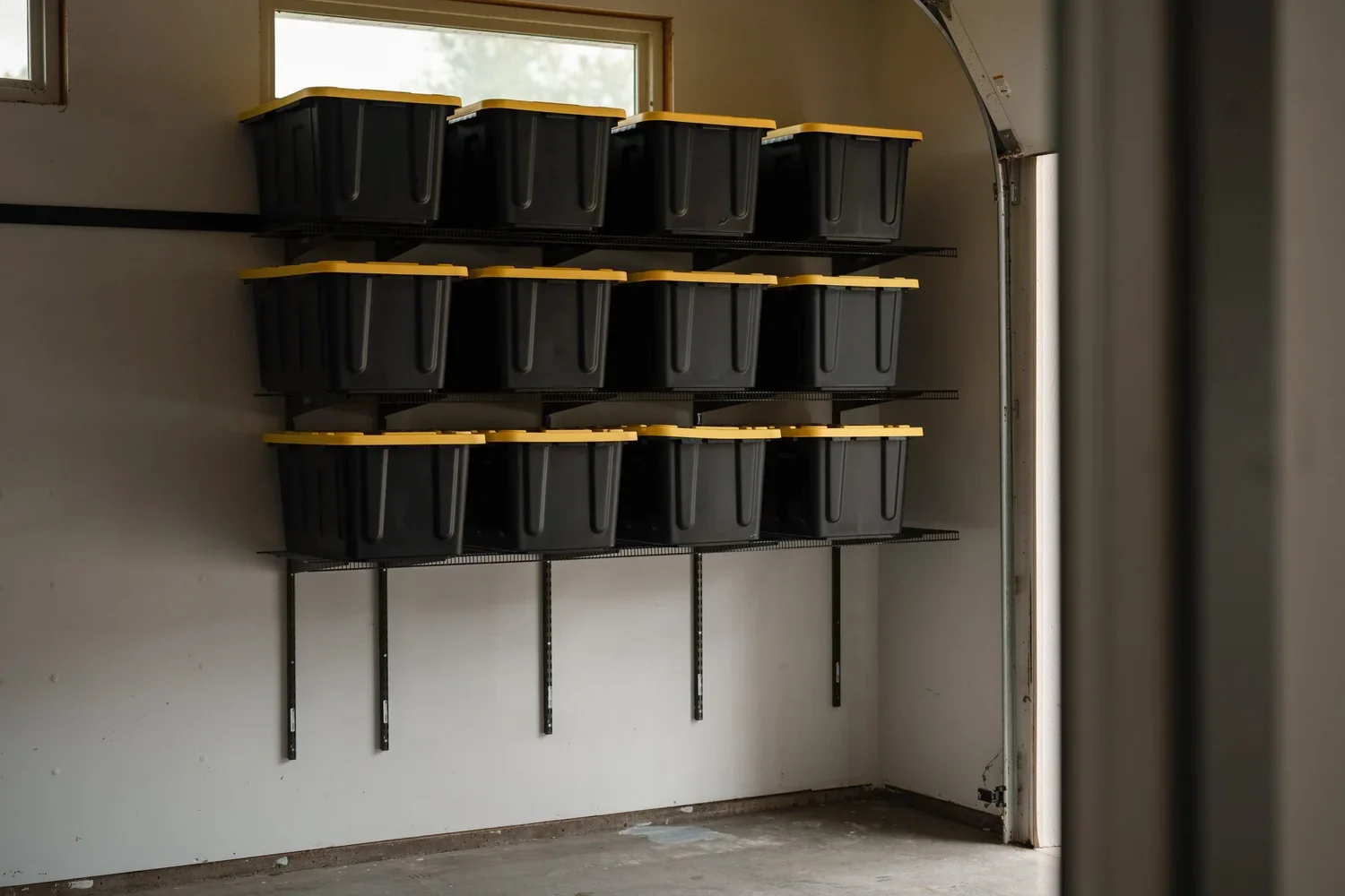 Black storage bins with yellow lids on three gunmetal shelves below a garage window