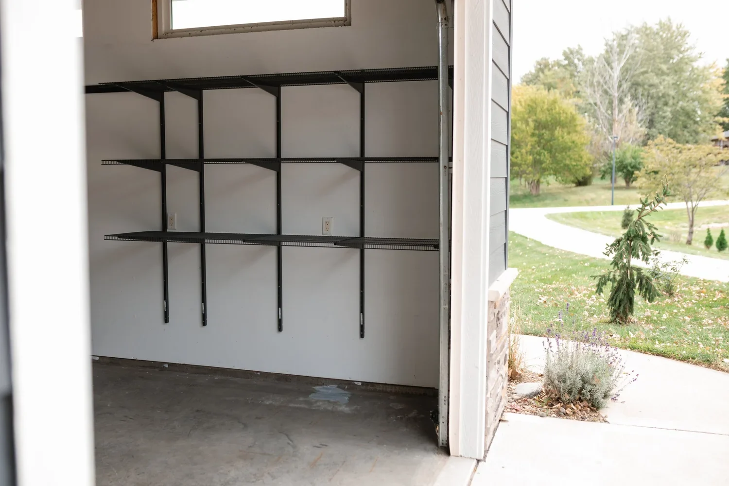 Empty gunmetal shelves on wall rails beneath a garage window, beside an open door