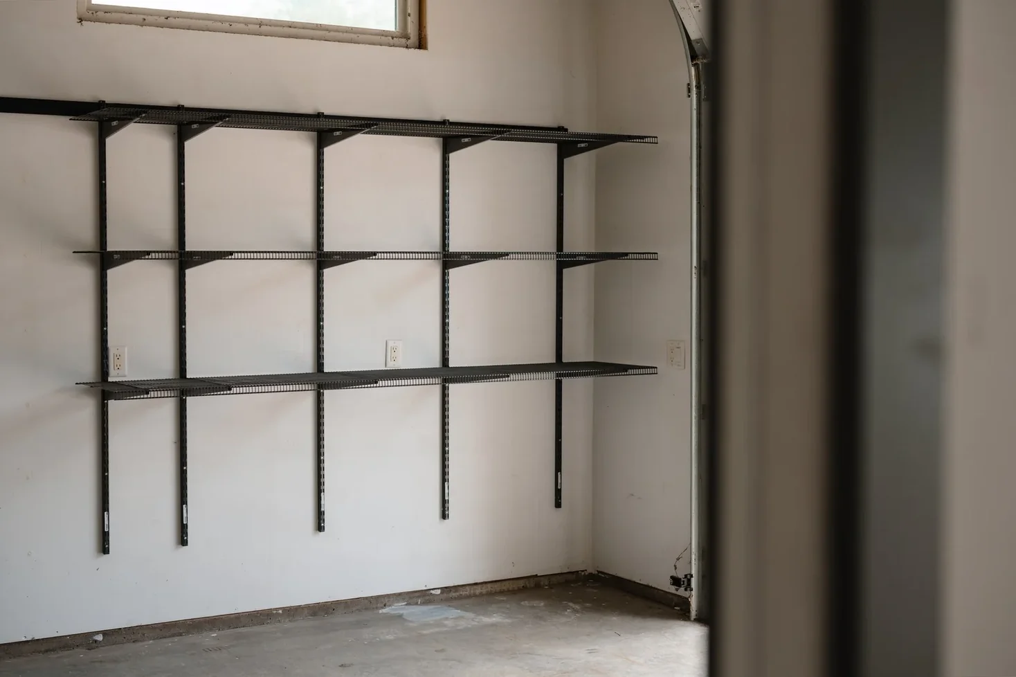 Empty gunmetal shelves on garage wall rails, beside the edge of the garage door