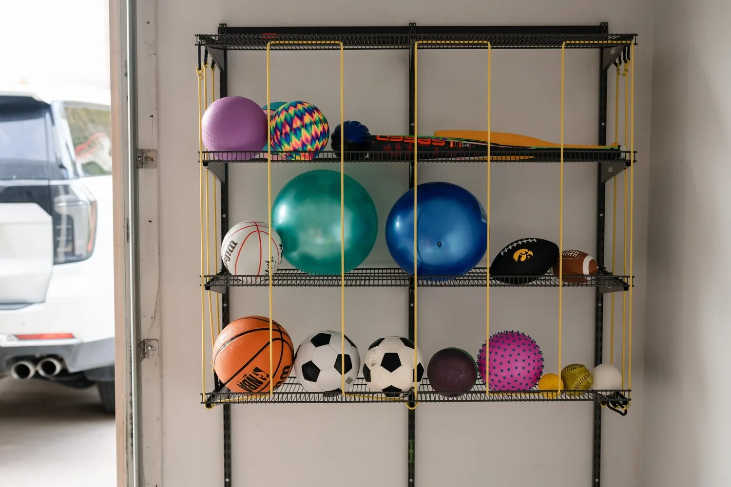 A ball rack filled with sports balls on gunmetal garage rails, next to a parked car