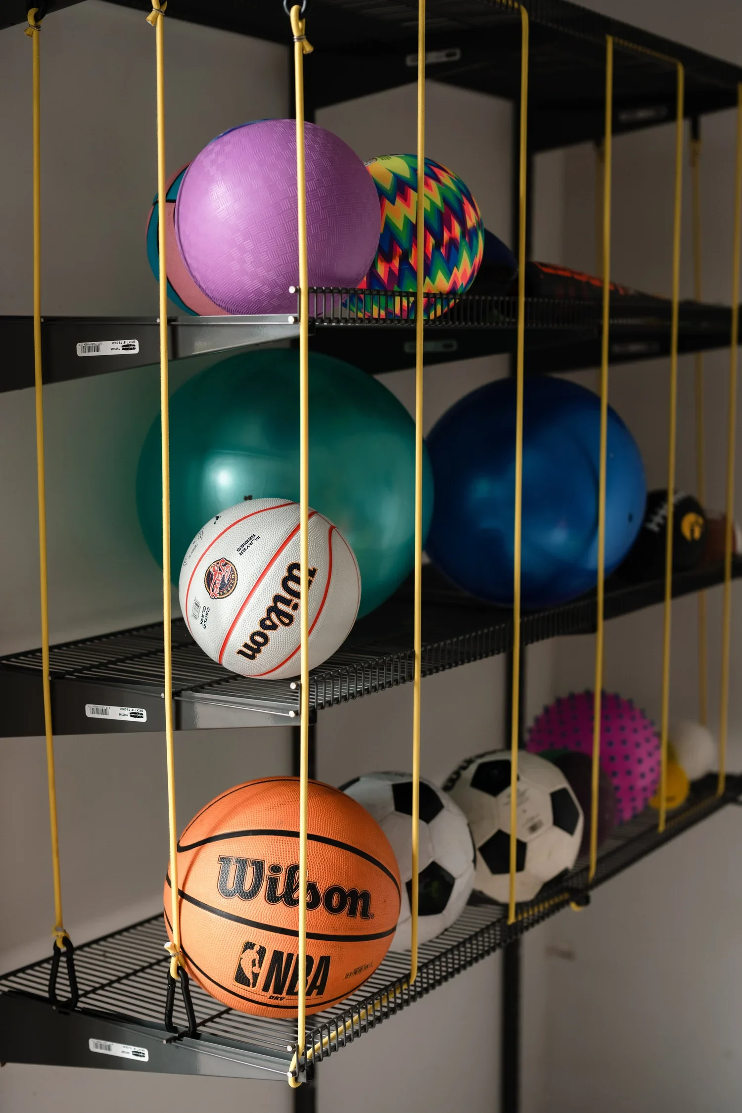 Close view of basketballs and soccer balls held behind yellow bungee cords on a garage ball rack