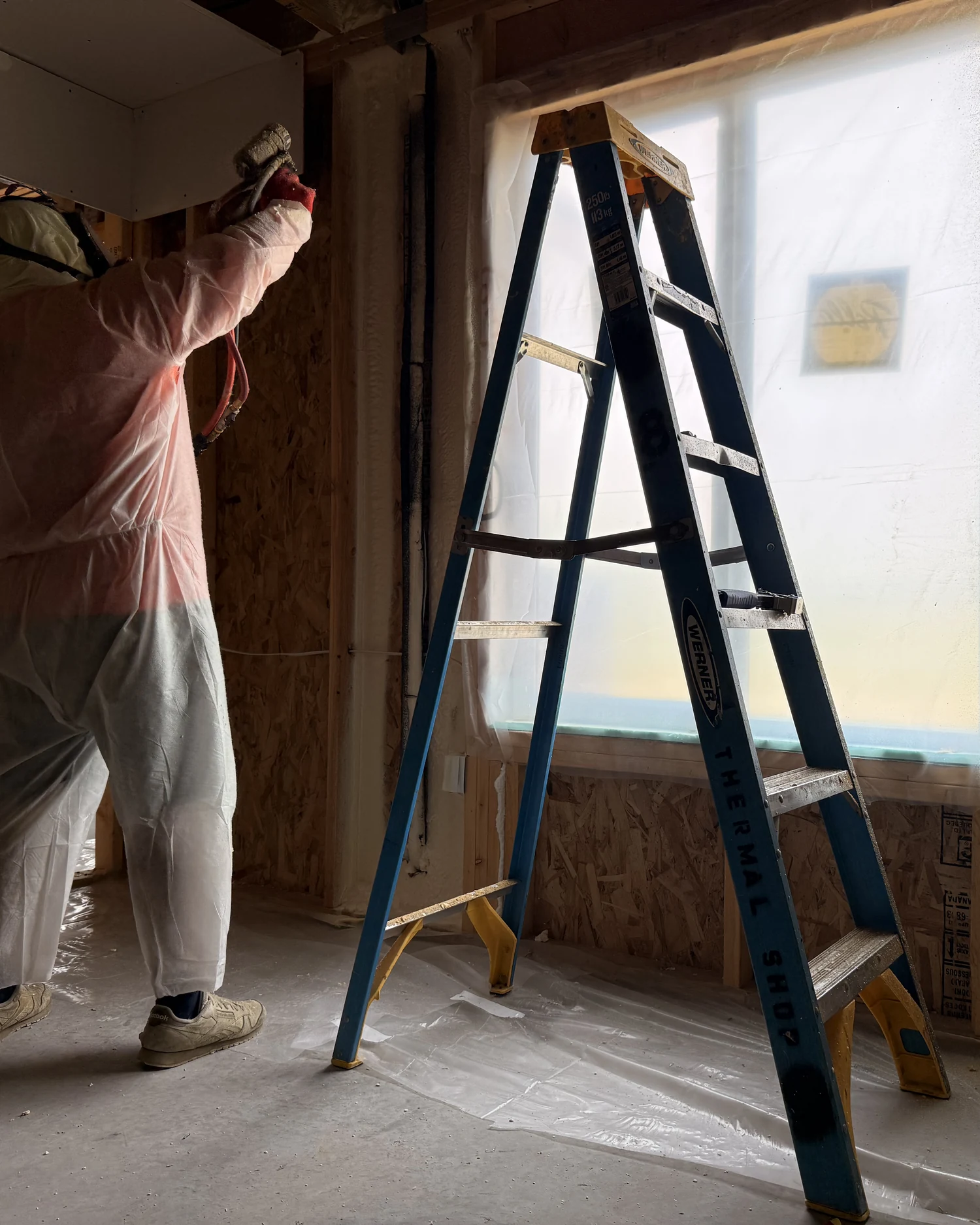 Installer in a protective suit spraying foam into open wall cavities, with a ladder and a plastic-covered window beside him